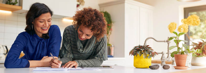 Two women enjoying working on details for their house, recording information on paper and a tablet at the kitchen counter